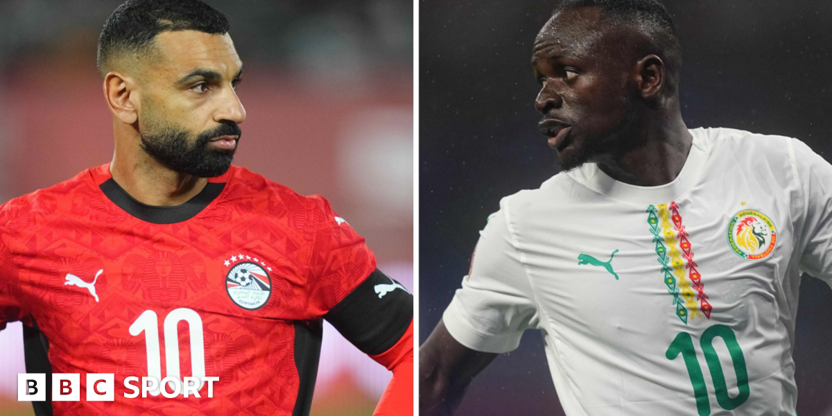 Mohamed Salah, wearing a red Egypt shirt with white 10 on the chest and a black captain's armband on his right bicep is seen looking to the right with his hands on his hips while Sadio Mane, wearing a white Senegal shirt with green, yellow and red detail and a green 10 on the chest, turns his head to the left