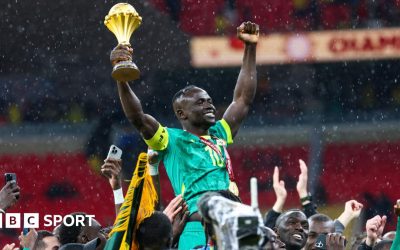 Brahim Diaz in all-white Morocco kit is seen on the left side of an image, chipping a penalty straight into the arms of Senegal goalkeeper Eduoard Mendy, all in blue on the right of the image, in the final of Afcon 2025. Mendy waits, bent slightly over, to collect the ball which is nearing him