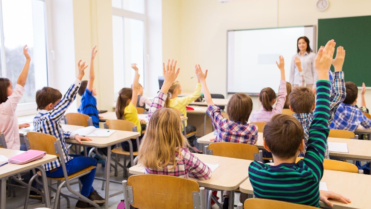 Mai mulți elevi, care stau în bănci, într-o sală de clasă, sunt cu mâinile ridicate