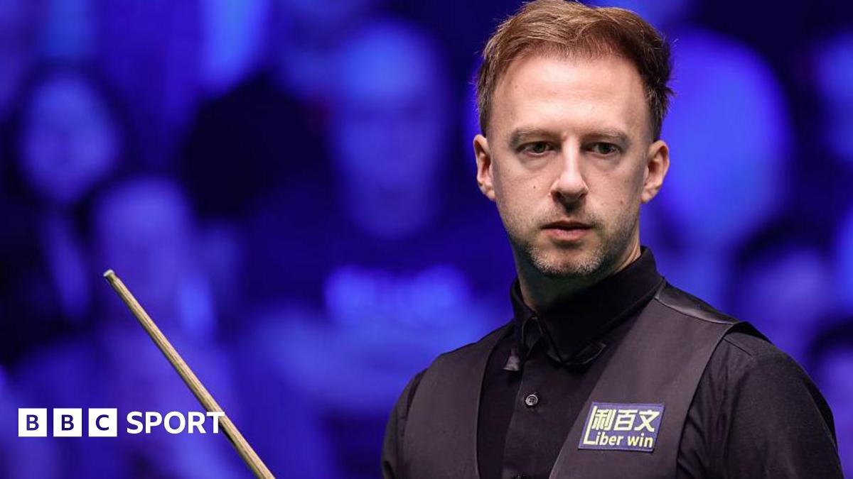 Judd Trump in black shirt and waistcoat carrying a snooker cue