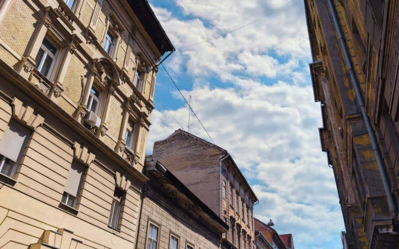 A budapesti hagyományos európai építészet felfelé ívelő látványa, amely elegáns, de időjárásnak kitett történelmi épületeket mutat be részletes homlokzatokkal és díszítőelemekkel. A keskeny utca perspektívája drámai keretet terem